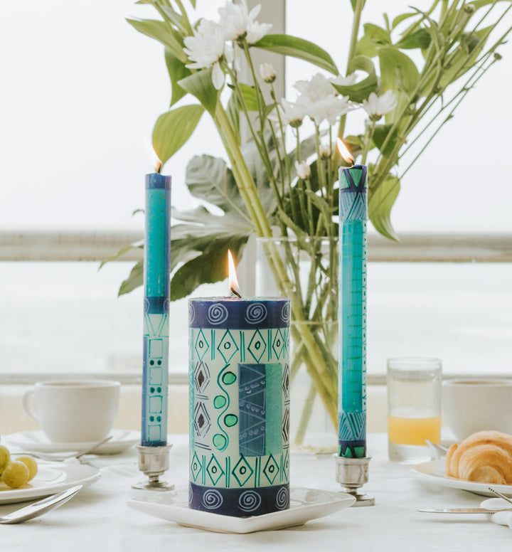 Lifestyle photo of Blue & Green tapers in pewter candle holders, with a 3x6 pillar.  On a breakfast table with some flowers in the background.  Fresh & bright!