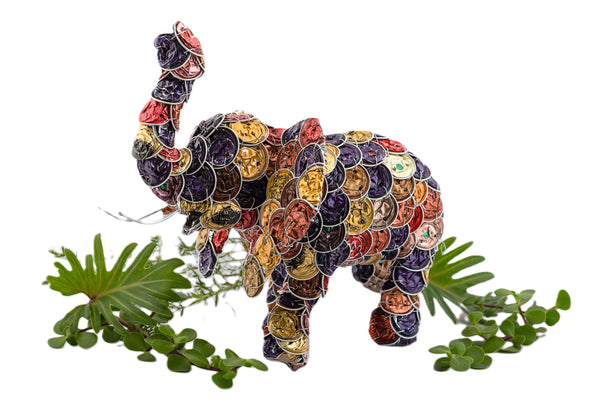 Wonderful elephant made from upcycled coffee pods in colors brown, gold, rust-red, and mauve.  Truck up for good luck!  The elephant is surrounded by some leaves and greenery.