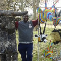 The artist, Godfrey at a craft fair in South Africa. He's standing in between various wired animal heads he created. From left to right: silver wire water buffalo head, silver wired Greater Kudu (horned antelope), other wire animal heads on the right side are colorful.