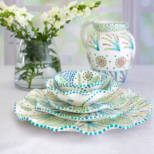 Life style photo of a stunning ceramic  plate collection.  White background with small floral design in blue & turquoise with touches of red and lime green. Stacked is a large ceramic dinner plate, dinner ceramic plate and side ceramic plate with matching ceramic salad bowl and ramekin.  Matching water jug in background. Beautiful!!