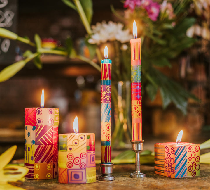 Lifestyle photo of the Carousel candle.  Pillars, tapers and a cube are all lite with some flowers in the background.   Soft and colorful!