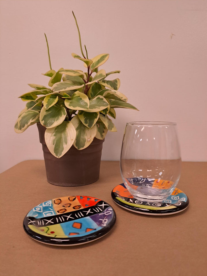 Two multicolor ethnic round ceramic coasters, hand painted and full of African color and designs. One showing how nicely a glass sits on the coaster.  Fair trade.