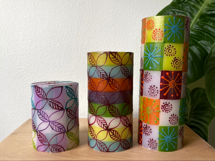 Magic Garden collection pillar candles; 3" x 4' in hues of purple with floral designs as overlay, 3" x 6" pillar candle with olive, burnt orange, cream, light blue and grey stripes with floral overlay,  and 3" x 8" pillar candle with same background colors in square, with funky flowers overlay.  All hand painted in South Africa.